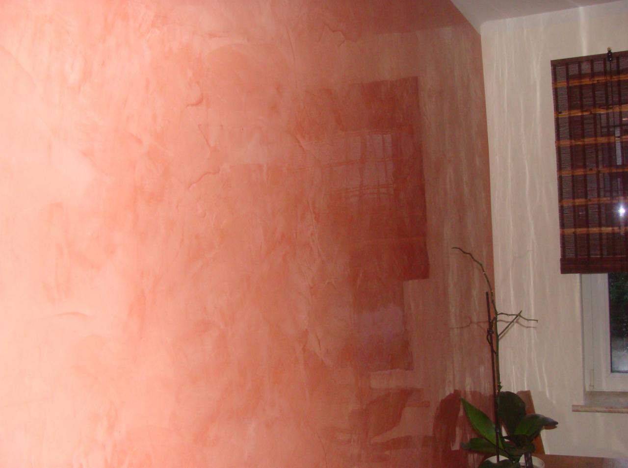 Ściana z efektem dekoracyjnym w odcieniach koralu i pomarańczy. Widoczna faktura gładzi z subtelnym połyskiem. W tle roślina doniczkowa i okno z bambusową roletą.