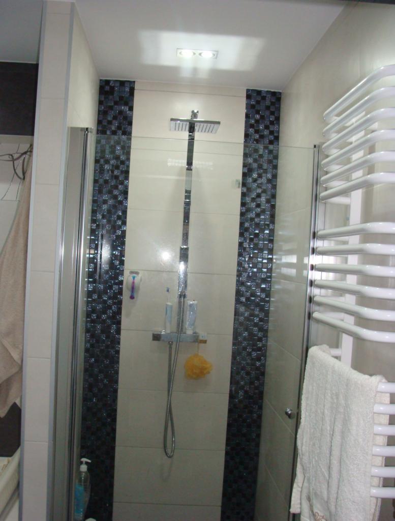 Wykończona kabina prysznicowa z mozaiką w odcieniach czerni i bieli, jasnymi płytkami i szklanymi drzwiami. Widoczny grzejnik drabinkowy z ręcznikiem.