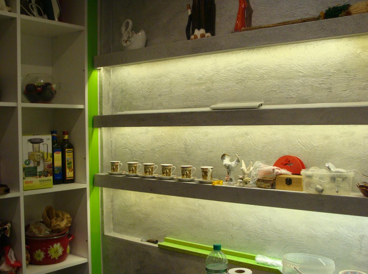 Ściana z dekoracyjną fakturą i półkami podświetlonymi ledami, na których stoją ozdoby i naczynia. Po lewej stronie regał z przedmiotami.