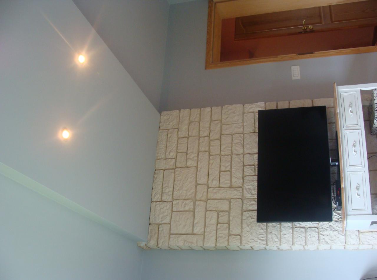 Ściana z dekoracyjnej cegły z zamontowanym telewizorem, oświetlona punktowo, z widoczną szafką w stylu vintage. Świeżo pomalowane ściany w odcieniach szarości.