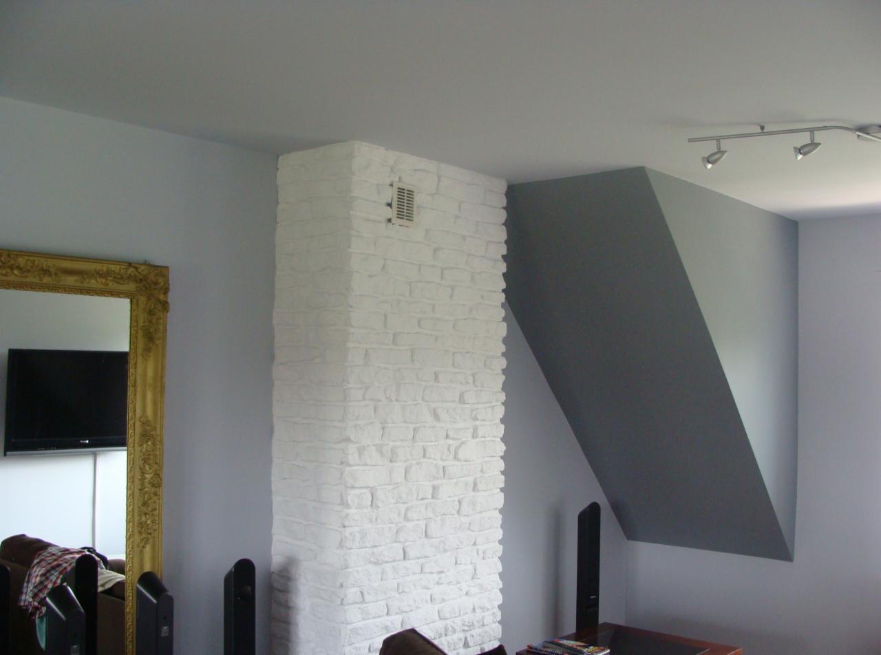 Pomieszczenie z białym, ceglanym filarem i szarą ścianą pod skosem. W tle złote lustro i fragment sofy, nowoczesne oświetlenie sufitowe.