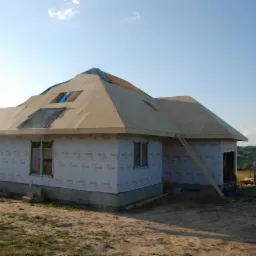 Surowy stan domu w trakcie budowy, z membraną ochronną Tyvek na ścianach i częściowo ukończonym dachem z widocznymi otworami, drabina oparta o ścianę, w tle pagórkowaty krajobraz.