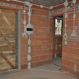 Wnętrze w stanie surowym z widoczną instalacją elektryczną w bruzdach, zabezpieczone deskami wejście do pomieszczenia, ściany z czerwonej cegły, widoczna skrzynka elektryczna i przewody.