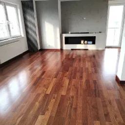 Salon z nową, błyszczącą podłogą z ciemnego drewna egzotycznego, widoczne okna z białymi ramami, grzejnik oraz dekoracyjny grzejnik pionowy, ściana z szarą, dekoracyjną fakturą i nowoczesny kominek.