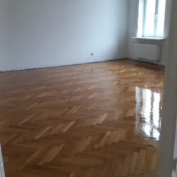 Parkiet ułożony w jodełkę, świeżo po cyklinowaniu i lakierowaniu, z widocznym połyskiem, w przestronnym pokoju z wysokim sufitem i ozdobnym gzymsem.