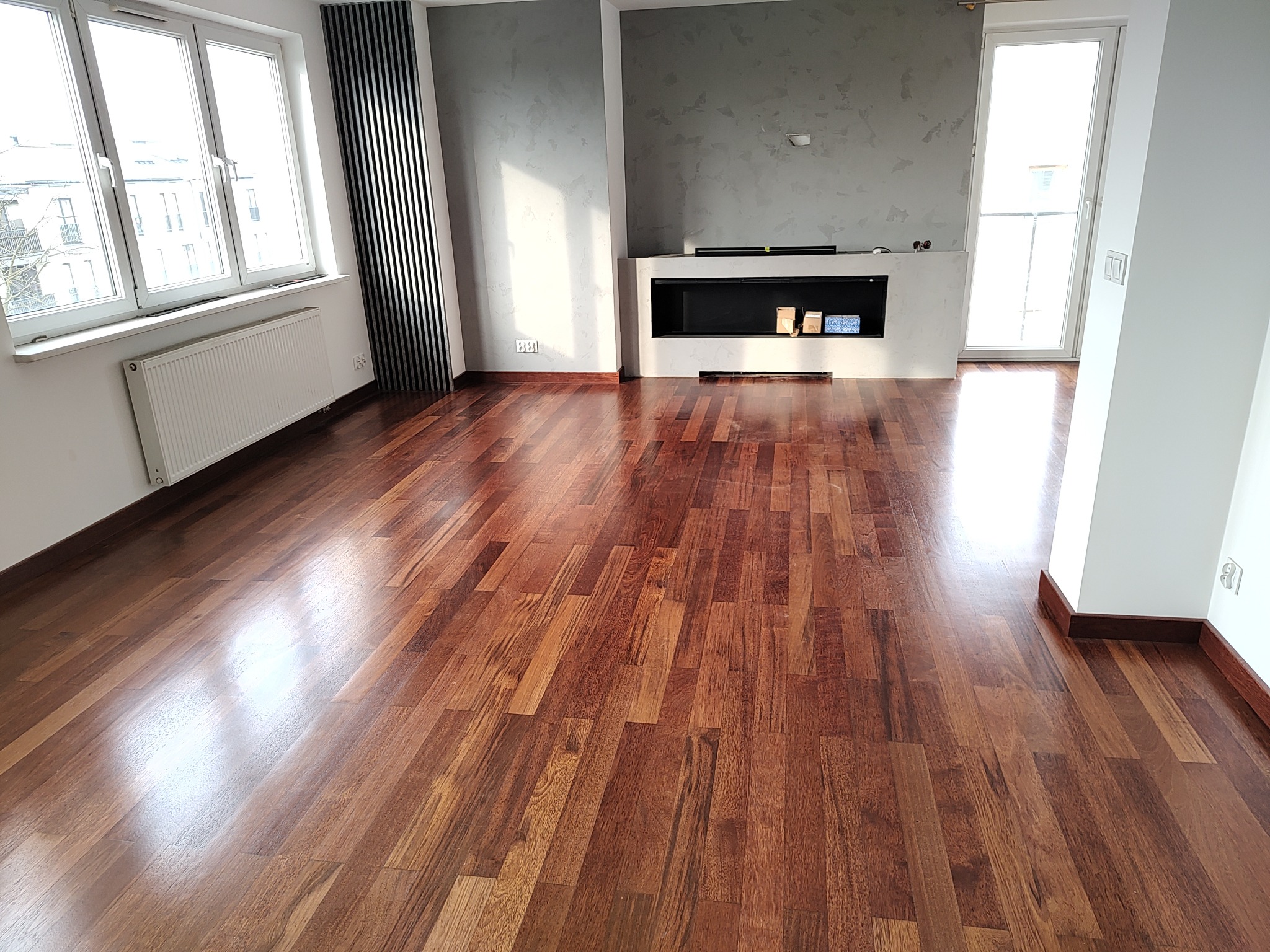Salon z nową, błyszczącą podłogą z ciemnego drewna egzotycznego, widoczne okna z białymi ramami, grzejnik oraz dekoracyjny grzejnik pionowy, ściana z szarą, dekoracyjną fakturą i nowoczesny kominek.