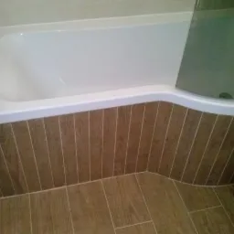 Biała wanna z obudową z płytek imitujących drewno, pasujących do płytek podłogowych, kabina prysznicowa częściowo widoczna po prawej stronie.