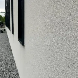 Ściana budynku z białą elewacją o wyraźnej fakturze, z czarnymi obramowaniami okien, widok z dołu, obok chodnik z szarym żwirem.