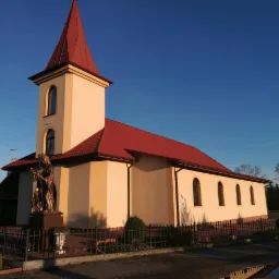 Kościół z beżową elewacją i czerwoną dachówką, widoczny pomnik przed budynkiem, otoczony metalowym ogrodzeniem i zielenią, na tle błękitnego nieba.