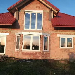 Dom w trakcie budowy z zamontowanymi białymi oknami, widoczne odbicie nieba i drzew w szybach, czerwona dachówka, surowa cegła na ścianach.