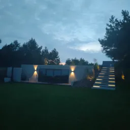 Nowoczesny ogród z betonowym budynkiem wkomponowanym w skarpę, oświetlonym schodami i trawnikiem, uchwycony o zmierzchu.