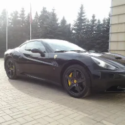 Czarny Ferrari California z żółtymi zaciskami hamulcowymi na tle budynku i drzew. Auto zaparkowane na kostce brukowej, widoczne detale karoserii.