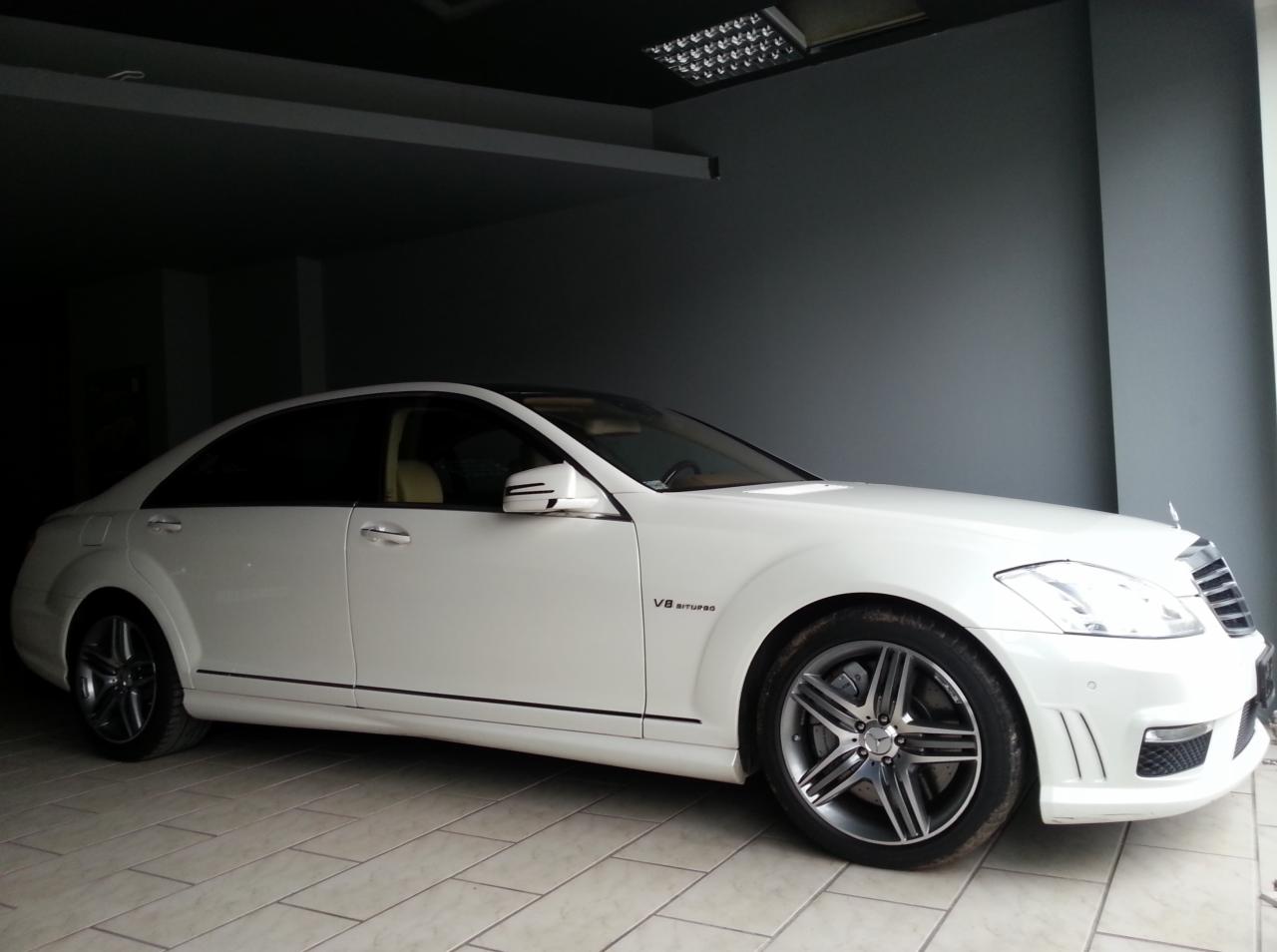 Biały Mercedes-Benz S63 AMG V8 Biturbo w garażu, z widocznymi detalami tuningu, takimi jak felgi i emblematy. Auto zaparkowane na jasnej, kafelkowej podłodze.