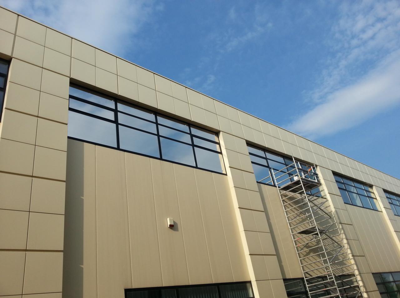 Elewacja budynku z oknami, na tle błękitnego nieba. Przy ścianie ustawione rusztowanie, na którym znajduje się osoba. Widok z dołu, podkreślający wysokość budynku.