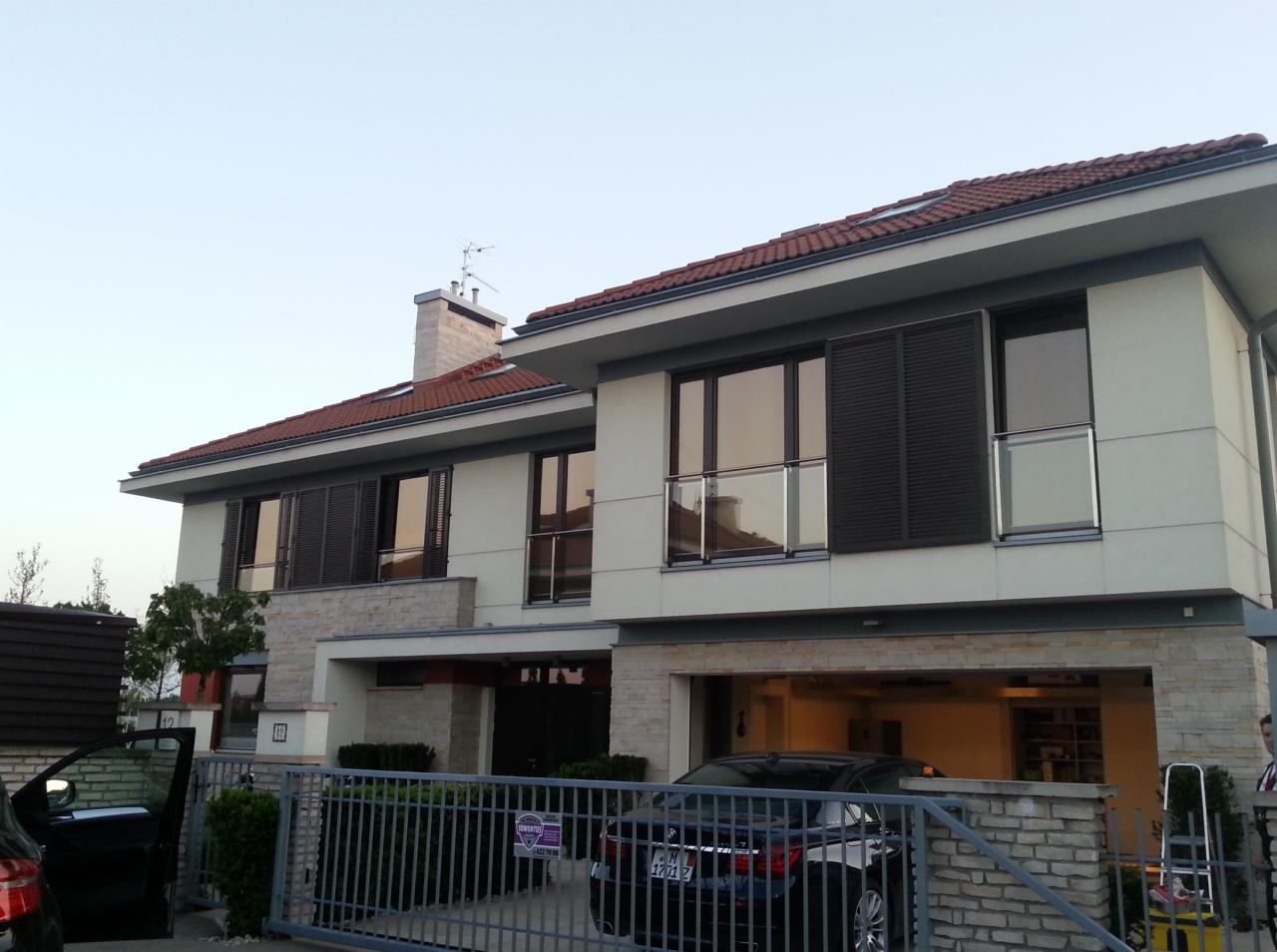 Dwupiętrowy dom z garażem i oknami z żaluzjami, widoczny zza ogrodzenia. Na podjeździe stoi czarny samochód.