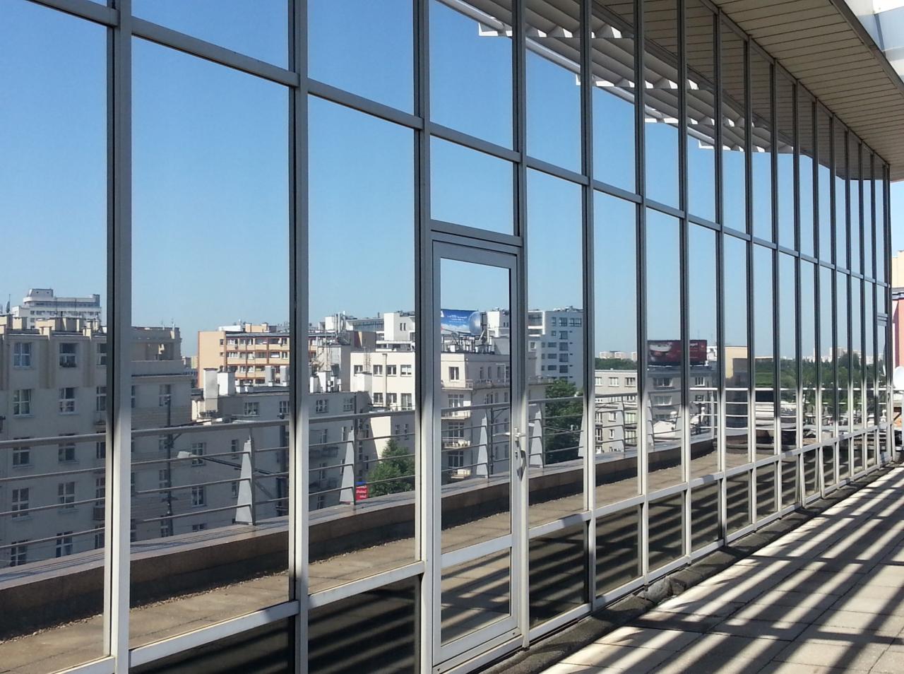 Szklana fasada budynku biurowego odbija panoramę miasta w słoneczny dzień. Widoczne regularne ramy okienne i cień na chodniku, podkreślające nowoczesny charakter architektury.