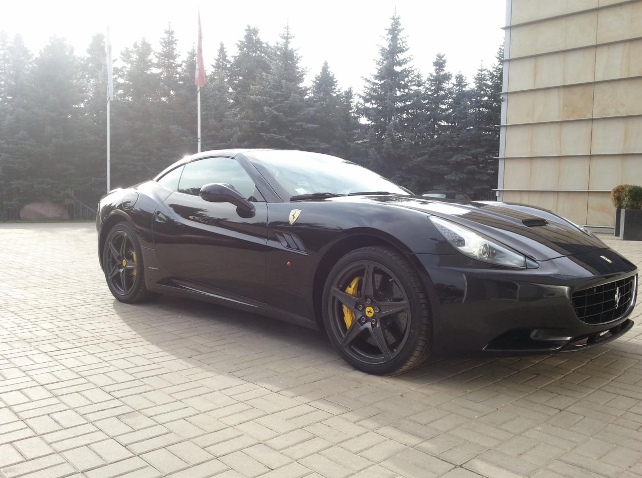 Czarny Ferrari California z żółtymi zaciskami hamulcowymi na tle budynku i drzew. Auto zaparkowane na kostce brukowej, widoczne detale karoserii.