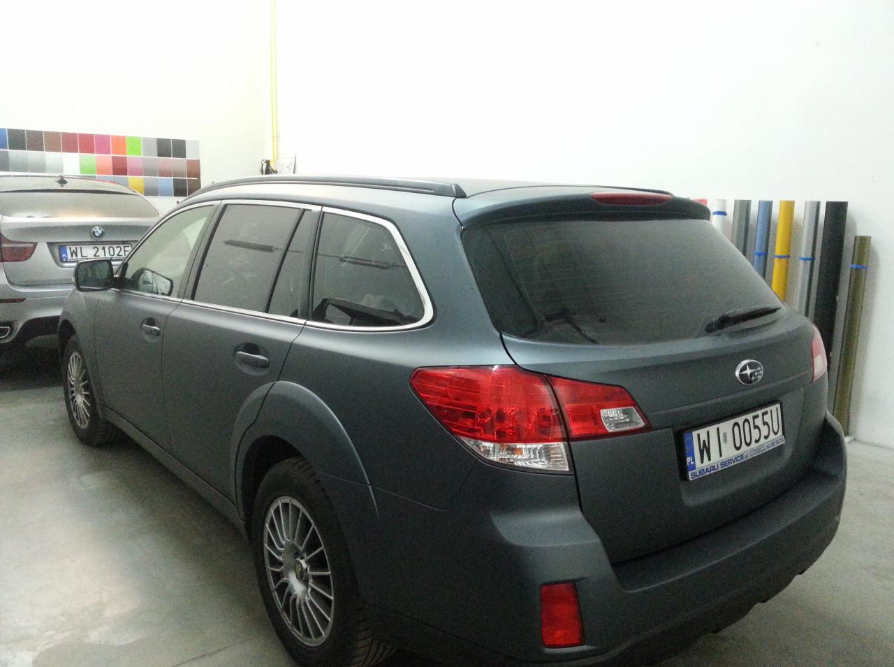 Szary samochód Subaru Outback po zmianie koloru folią matową, widoczne detale karoserii i tylne światła. W tle wzornik kolorów i rolki folii.