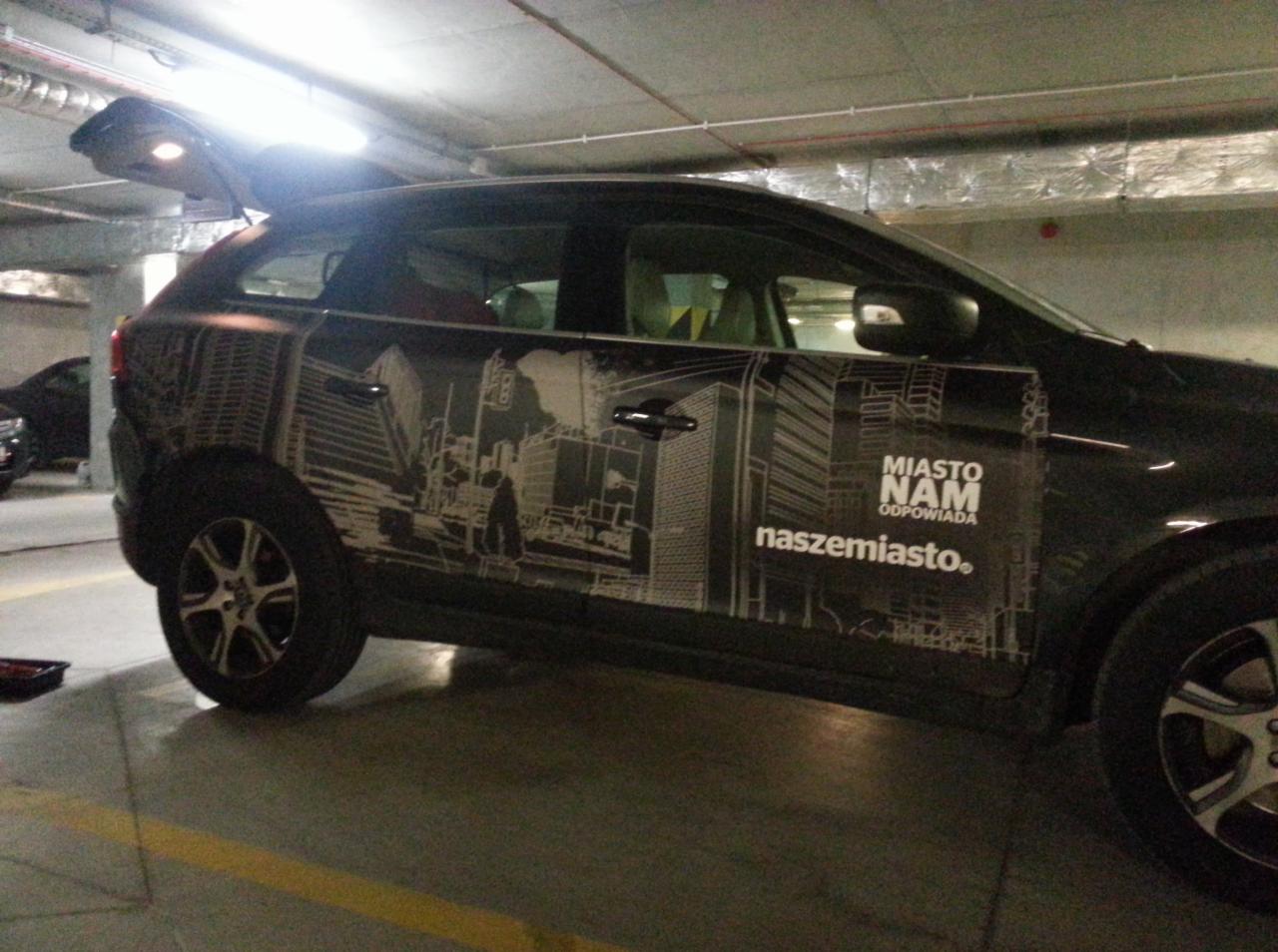 Ciemny samochód z grafiką miejską i napisem 'Miasto Nam Odpowiada' na boku, zaparkowany w garażu podziemnym z otwartą klapą bagażnika.