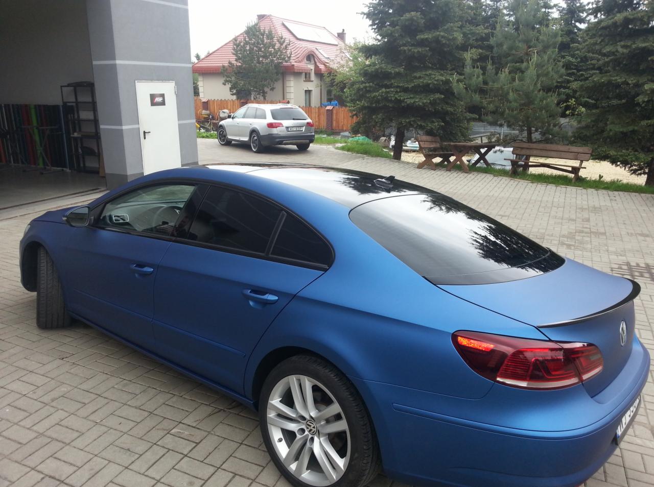 Niebieski Volkswagen CC po tuningu, matowa folia, czarny dach, spojler. W tle budynek i inny samochód. Widok z tyłu, lekko z boku, na kostce brukowej.