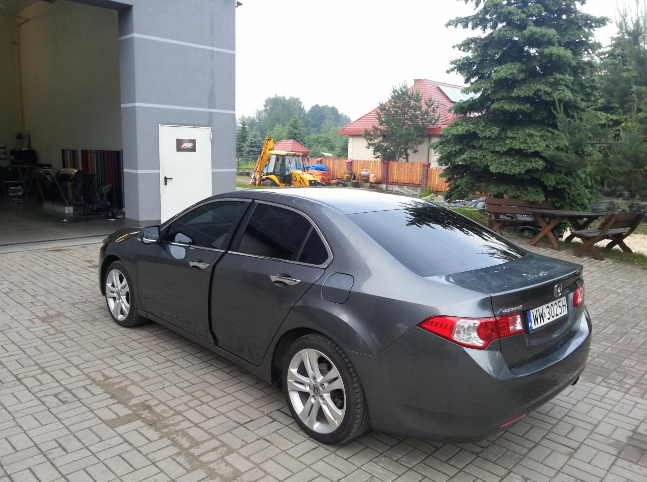 Przyciemnione szyby w szarym samochodzie Honda Accord, zaparkowanym przed budynkiem warsztatu z widocznymi rolkami folii i sprzętem budowlanym w tle.