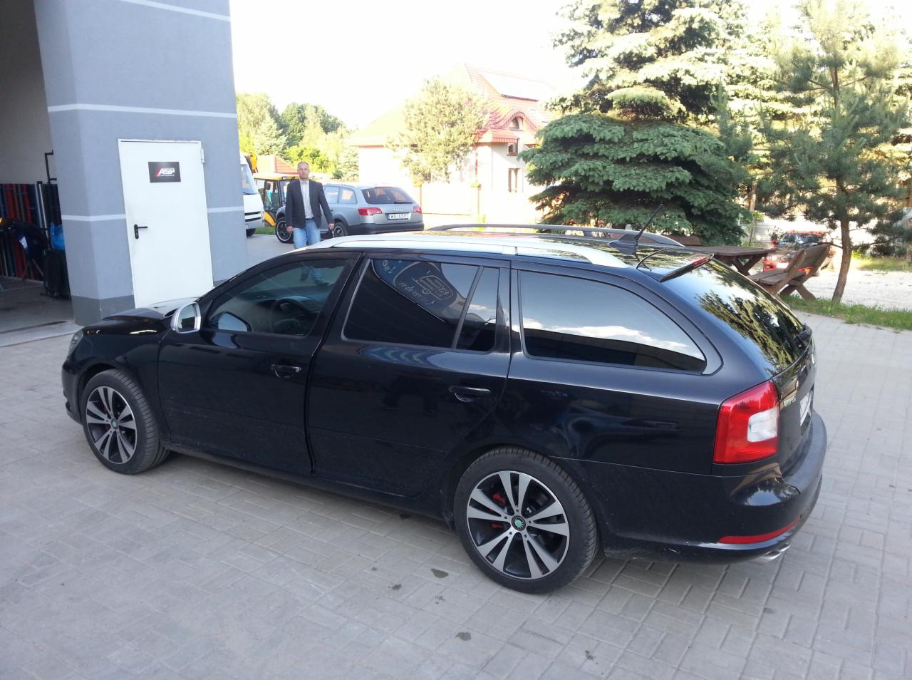 Czarna Skoda Octavia Combi z przyciemnionymi szybami i felgami aluminiowymi stoi na kostce brukowej przed budynkiem. Widoczny mężczyzna w tle.