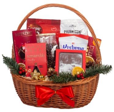 Wiklinowy kosz z czerwoną kokardą wypełniony słodyczami, kawą, suszonymi pomarańczami i dekoracjami świątecznymi, ułożony na białym tle.