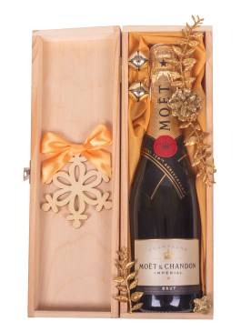 Otwarty drewniany kosz prezentowy z butelką szampana Moët & Chandon Imperial, złotymi ozdobami florystycznymi i drewnianą kokardą w kształcie śnieżynki, na żółtym jedwabnym tle.