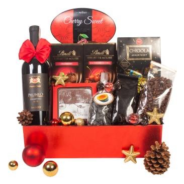 Czerwony, błyszczący kosz wypełniony butelką wina z czerwoną kokardą, czekoladami Lindt, kawą ziarnistą, pralinkami, ozdobami świątecznymi w postaci złotych gwiazdek i bombek oraz szyszkami.