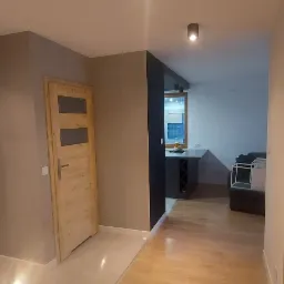 Wykończony korytarz z drewnianą podłogą i drzwiami, widok na salon z czarną ścianą akcentującą i nowoczesnym oświetleniem punktowym.