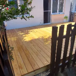 Świeżo położony taras drewniany z widocznymi puszkami z impregnatem Remmers i pędzlem malarskim przy ogrodzeniu, w tle budynek mieszkalny.