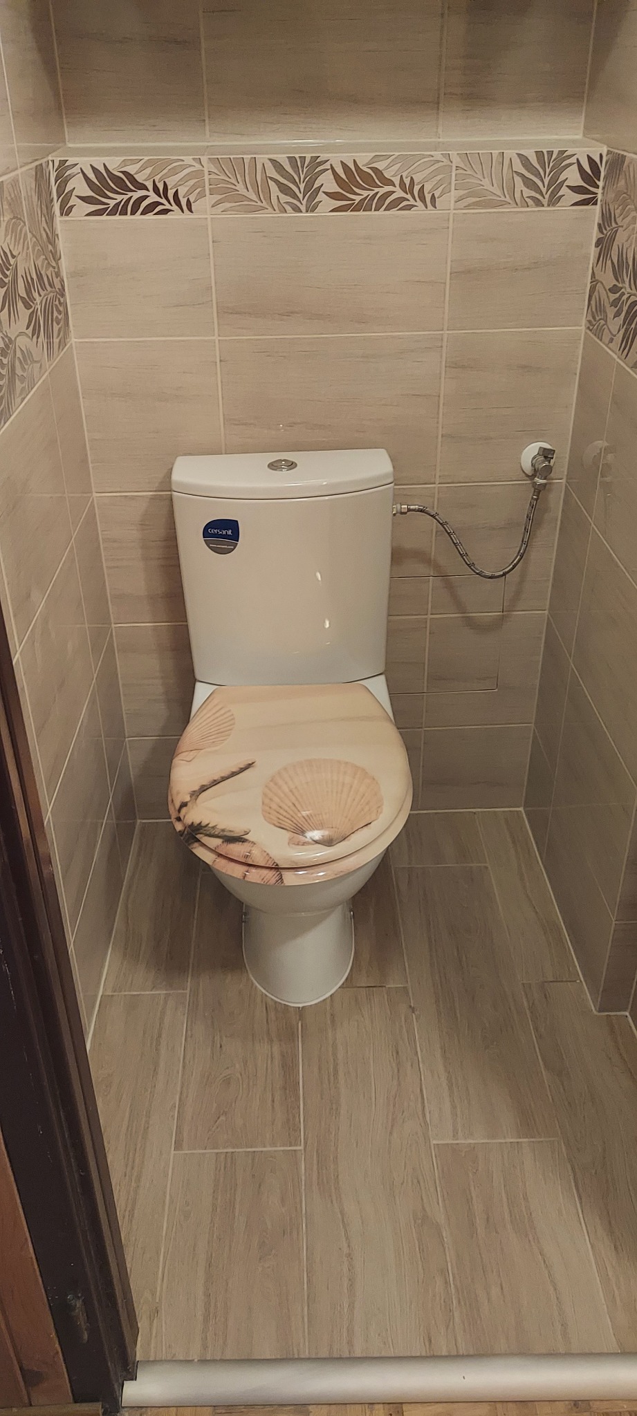Beżowa łazienka z toaletą z deską ozdobioną motywem muszli, płytki imitujące drewno na podłodze, dekoracyjny pas z motywem roślinnym na ścianach.