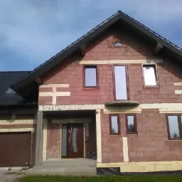 Dom w trakcie budowy z zamontowanymi oknami w brązowych ramach i częściowo ocieploną elewacją z czerwonej cegły.