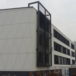 Nowoczesny budynek biurowy z białą fasadą ozdobioną trójkątnymi wzorami, czarnymi okiennicami i metalową konstrukcją osłaniającą okna, widoczny w pochmurny dzień.