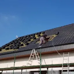 Dekarz montuje dachówkę na dachu domu jednorodzinnego, widoczne rusztowanie i drabina, część dachu jeszcze bez pokrycia z widoczną membraną dachową.