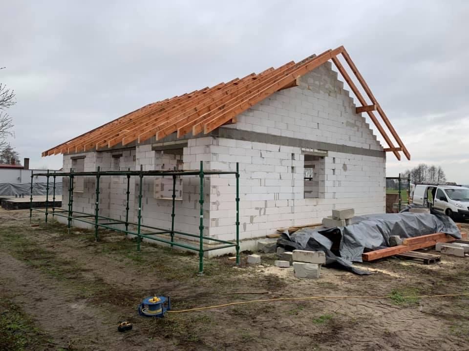 Budowa domu z białych bloczków: widoczne rusztowanie, więźba dachowa, materiały budowlane i częściowo wykończone ściany w pochmurny dzień.