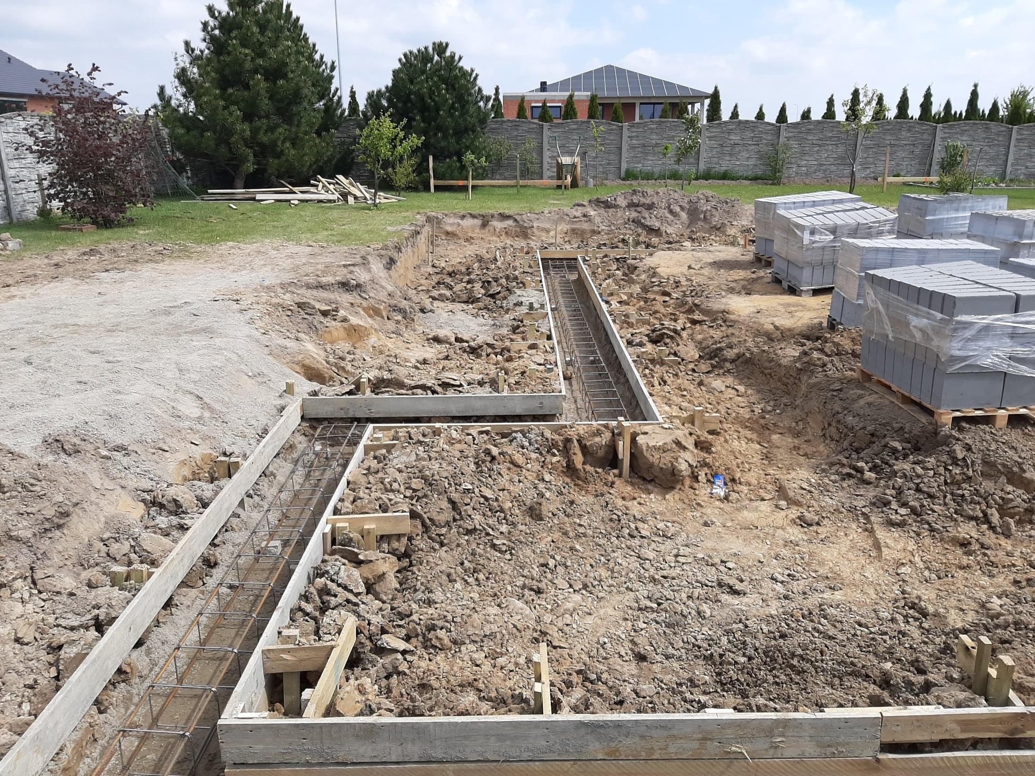 Przygotowanie fundamentów pod budowę domu: wykopane rowy z szalunkiem i zbrojeniem, teren częściowo wyrównany, w tle zapakowane bloczki betonowe i ogrodzenie.