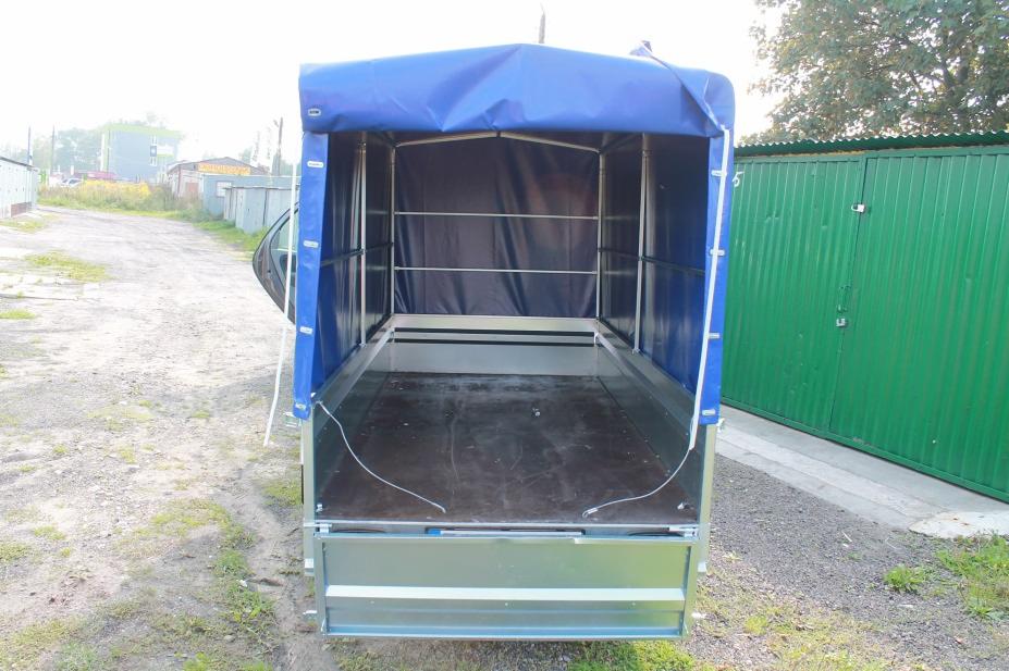 Pusta przyczepa transportowa z plandeką w kolorze niebieskim, widok z tyłu, na tle zielonego garażu blaszanego i drogi gruntowej.