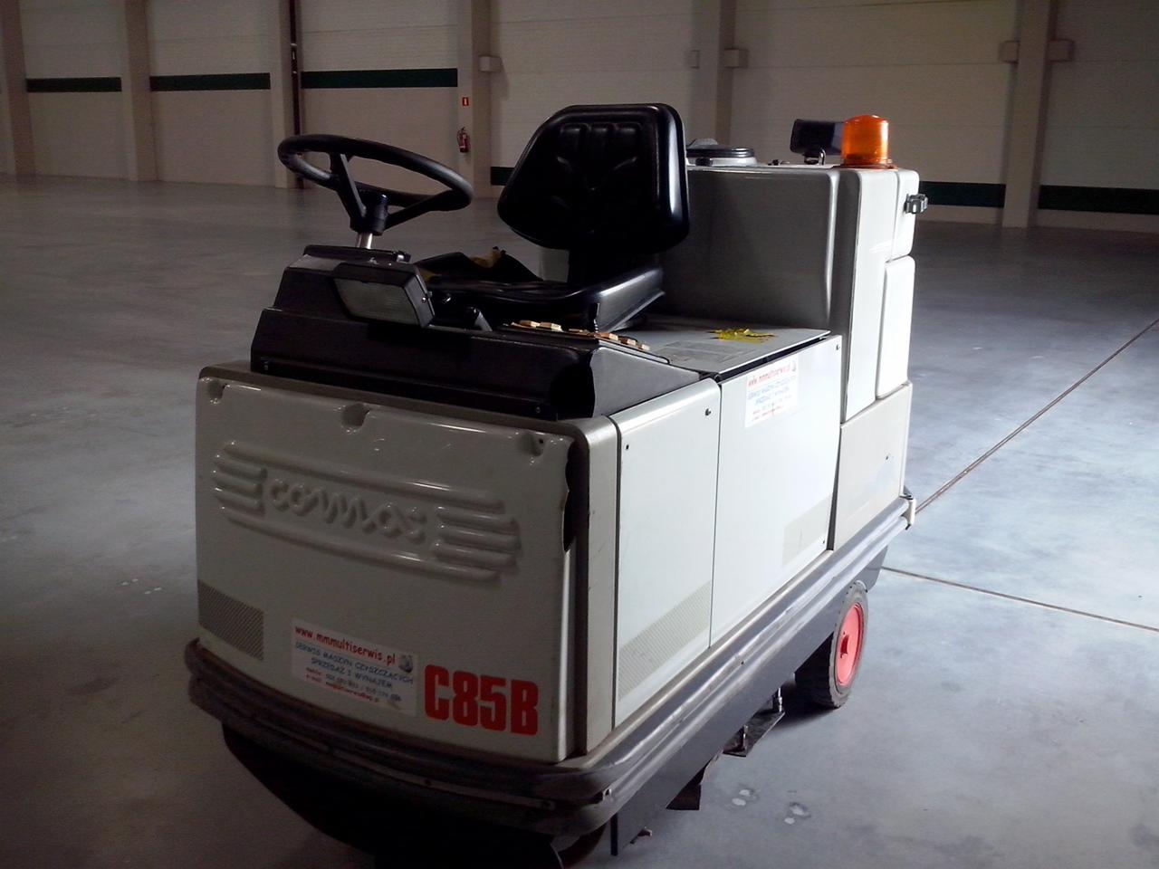 Szorowarka Comac C85B z widocznym logiem producenta, zaparkowana w hali magazynowej z betonową posadzką, oświetlona światłem dziennym.