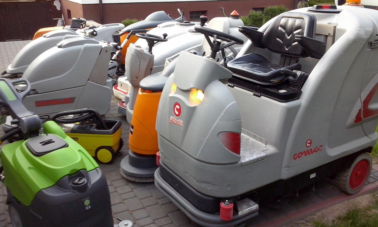 Różnokolorowe szorowarki do posadzek Comac Ultra 85B i ICA ustawione obok siebie na brukowanej powierzchni, w tle budynek.