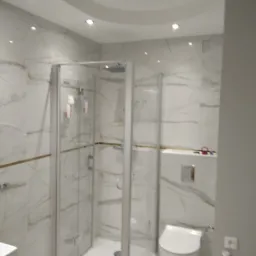 Nowoczesna łazienka z kabiną prysznicową typu walk-in, wykończona białymi płytkami imitującymi marmur z delikatnymi złotymi żyłkami, podwieszana toaleta, oświetlenie punktowe w suficie.