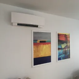 Biała klimatyzacja ścienna Daikin zamontowana w nowoczesnym salonie z abstrakcyjnymi obrazami w białych ramkach na białej ścianie.