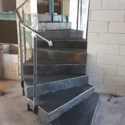 Metalowe schody z blachy ryflowanej z poręczą, zamontowane w surowym wnętrzu budynku w trakcie remontu, widoczne ślady prac budowlanych i fragmenty ścian z betonu komórkowego.