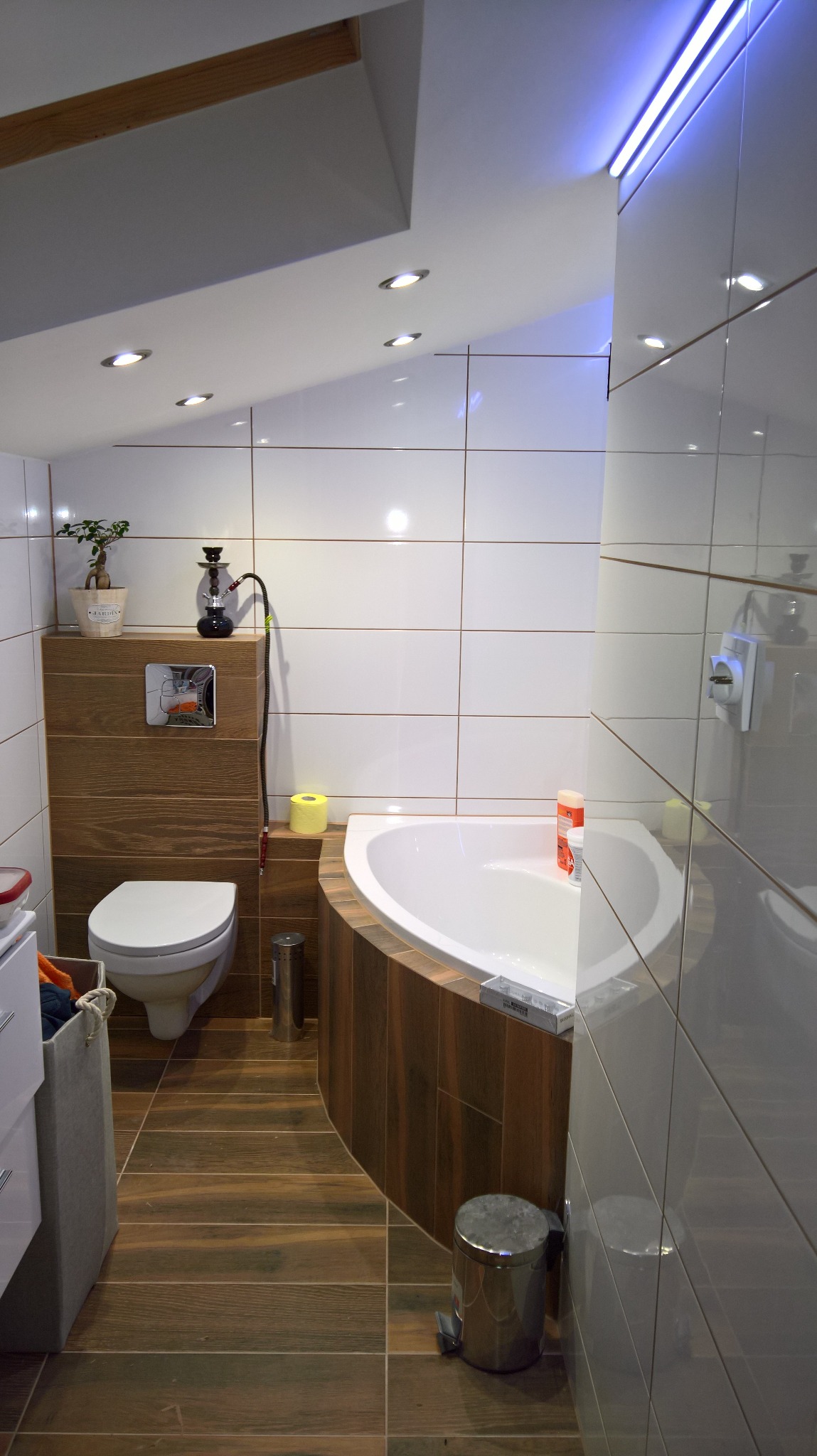 Nowoczesna łazienka z narożną wanną obudowaną drewnopodobnymi panelami, wiszącą toaletą z chromowanym przyciskiem spłukiwania, podwieszanym sufitem z oświetleniem punktowym i listwą LED,...