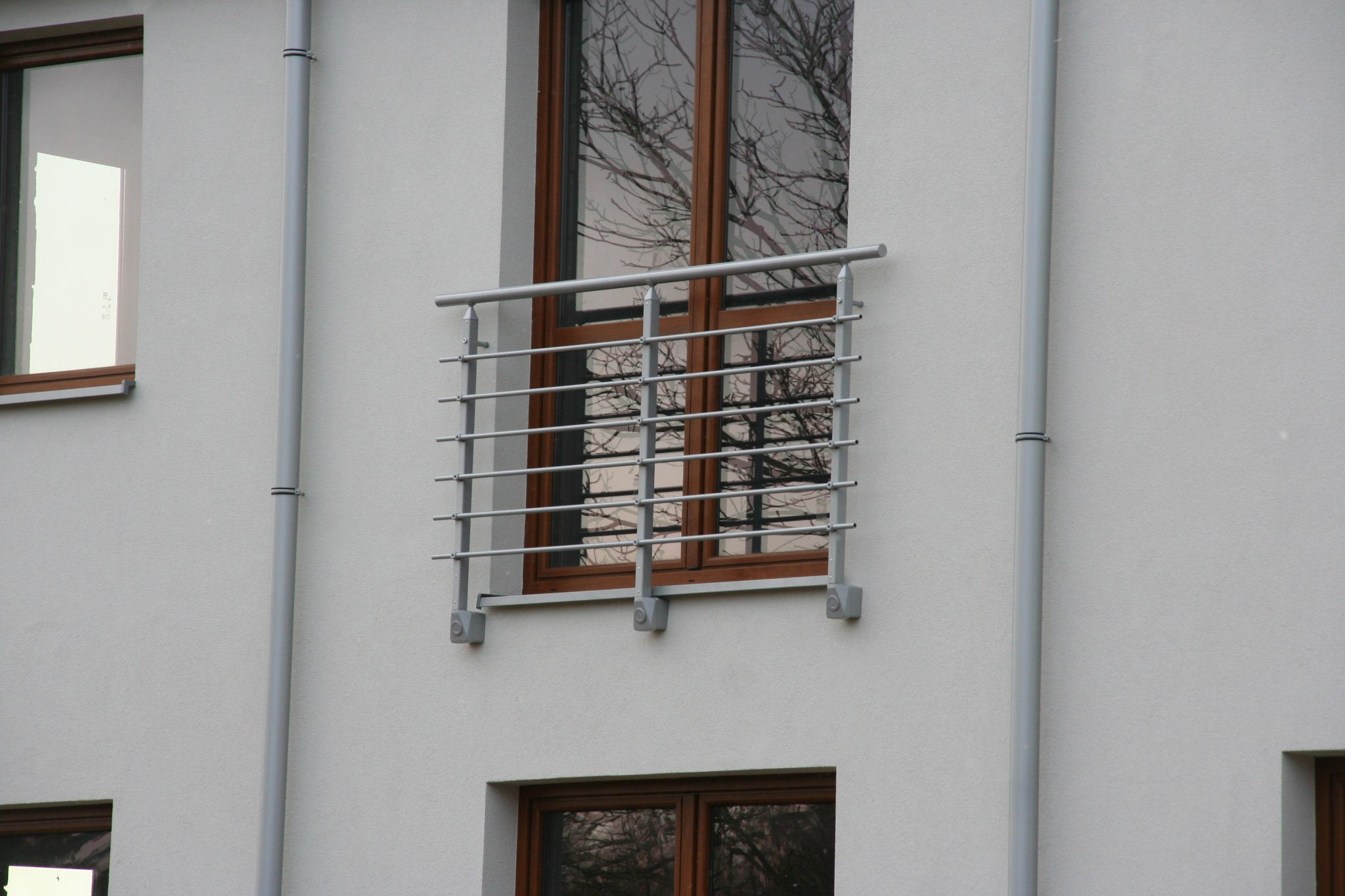 Elewacja budynku z oknami i balkonem zabezpieczonym metalową balustradą. W szybach okien odbijają się nagie gałęzie drzew.