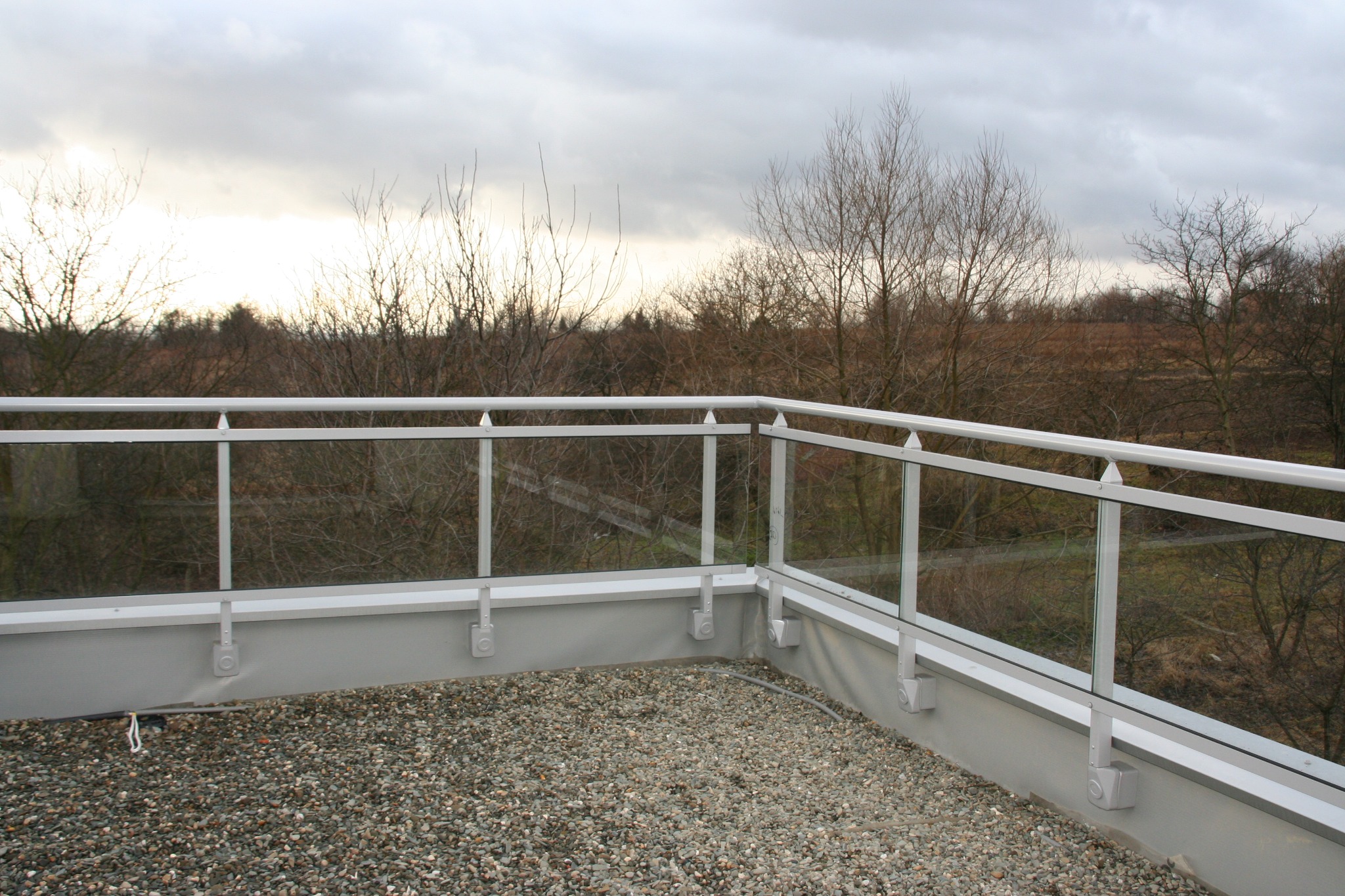 Wykończony taras na dachu z białymi balustradami z przezroczystym szkłem i wysypanym drobnym żwirem, widok na drzewa bez liści i pochmurne niebo.