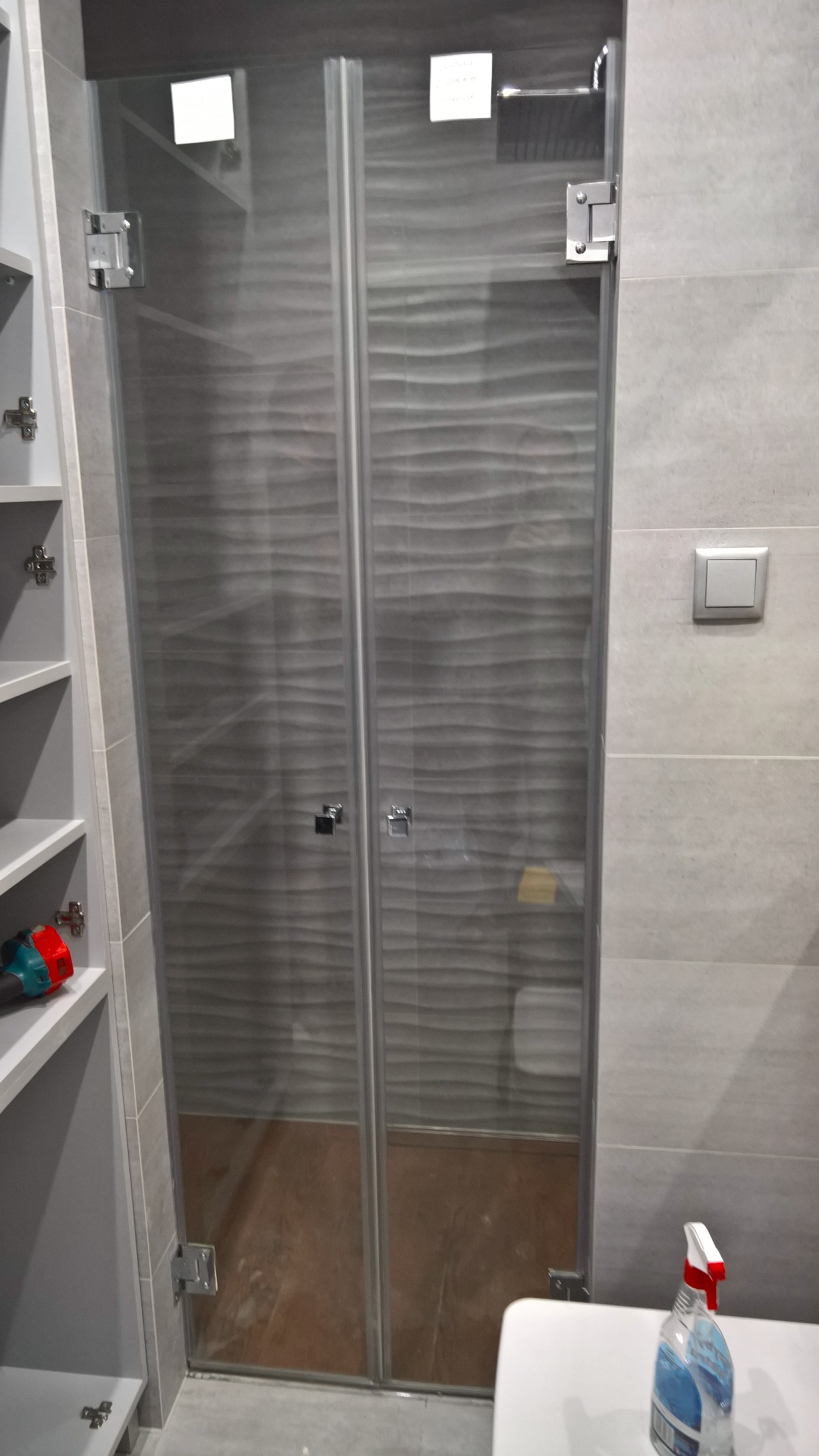 Szklana kabina prysznicowa z dwuskrzydłowymi drzwiami, zamontowana w łazience z szarymi płytkami, widoczne chromowane zawiasy i uchwyty, obok szafka z otwartymi półkami, na blacie butelka z płynem...