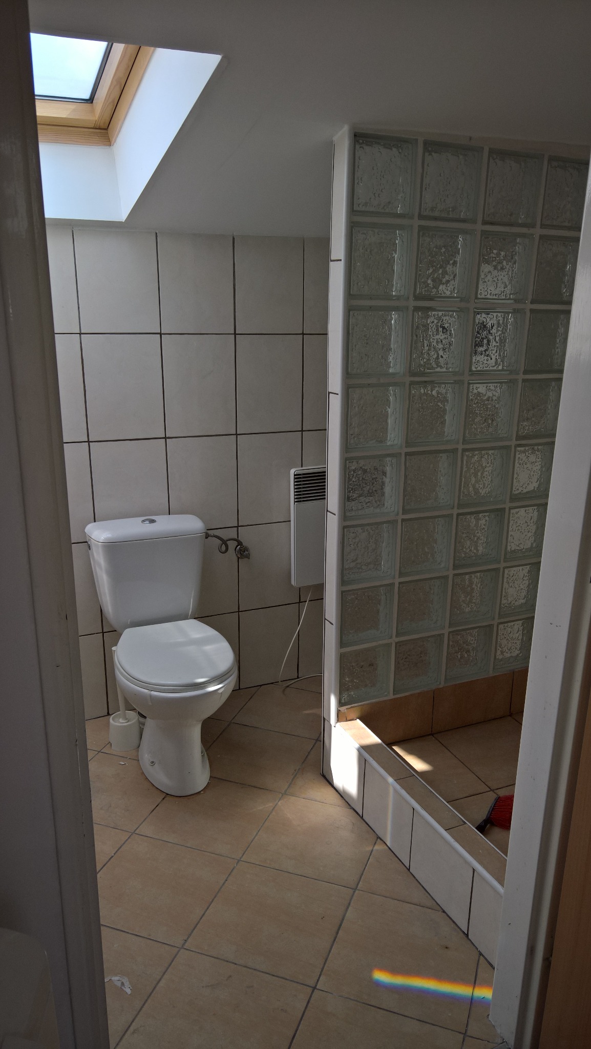 Wykończona łazienka z toaletą, grzejnikiem na ścianie i kabiną prysznicową z luksferów, oświetlona światłem słonecznym wpadającym przez okno dachowe.