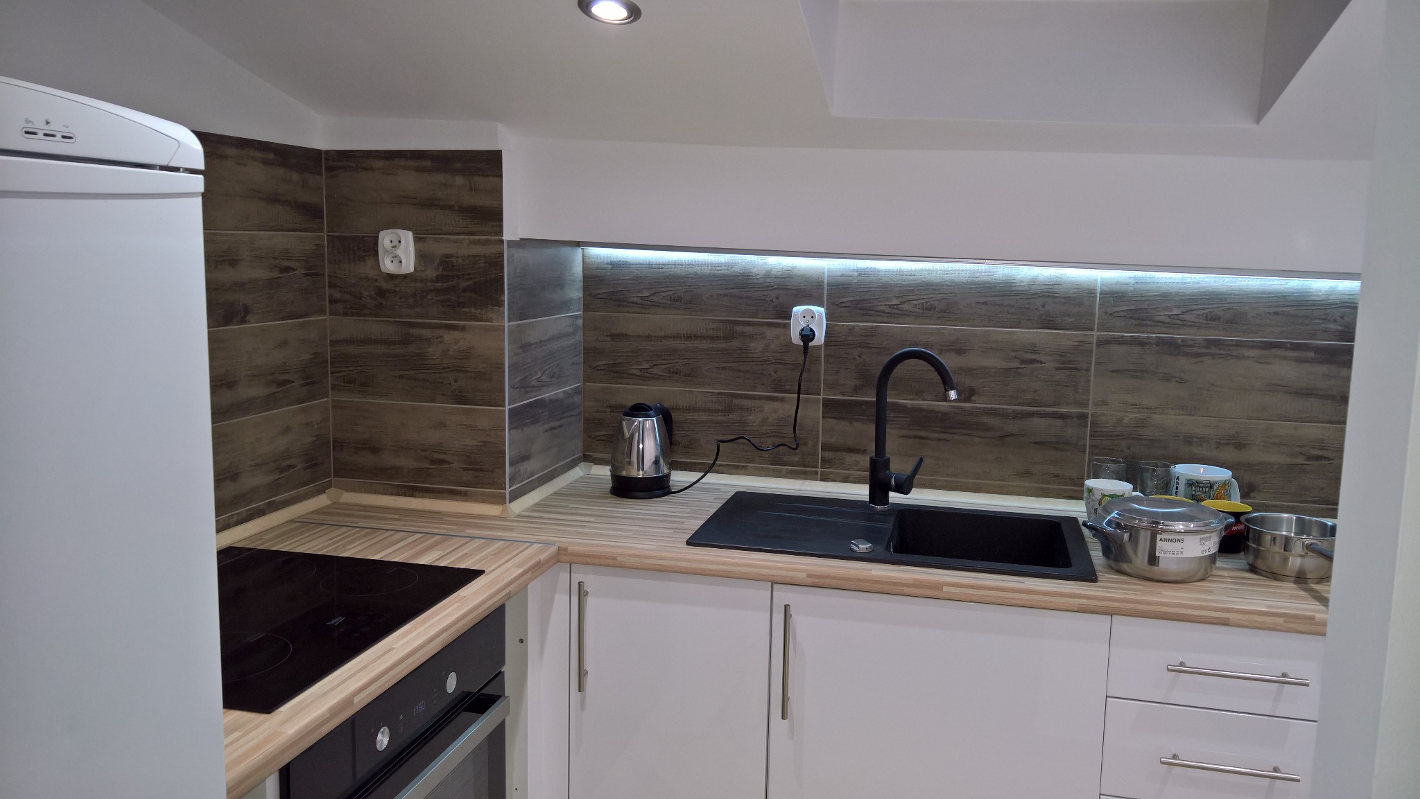 Nowoczesna kuchnia z białymi szafkami, czarnym zlewem i baterią, oraz płytkami imitującymi drewno na ścianie. Widoczne garnki i czajnik.