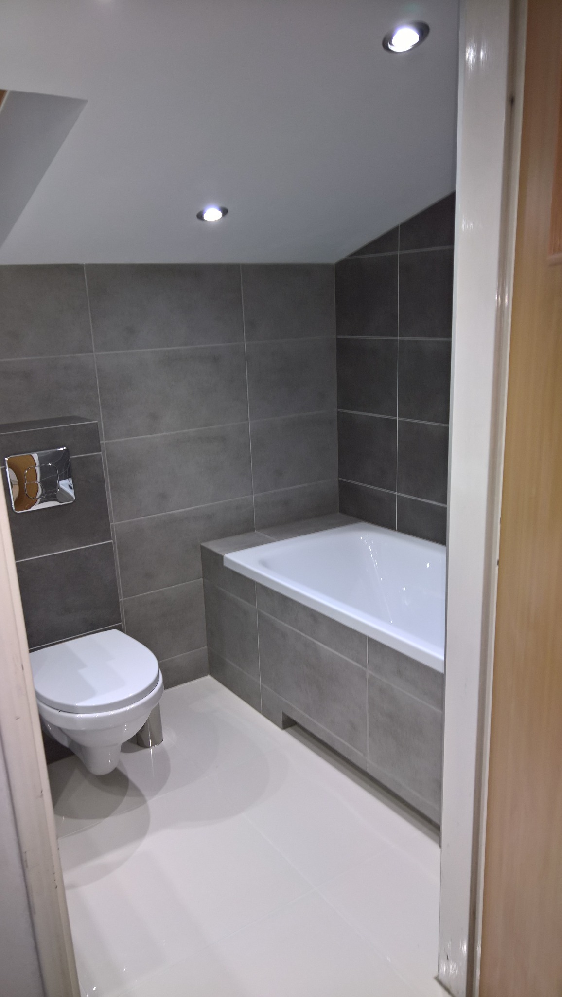 Nowoczesna łazienka z szarymi płytkami, białą wanną i toaletą, oświetlona punktowymi lampami sufitowymi.